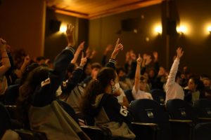 Crianças num cinema com as mãos levantadas apontando pra cima.