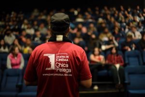 Homem de costas, em pé, num palco na frente de pessoas nas cadeiras de cinema. Veste uma camisa vermelha com a logo da Mostra de Cinema Chica Pelega.