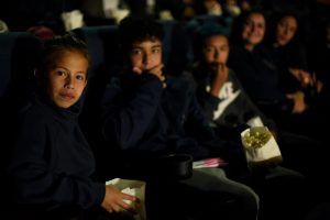 Cinco pessoas olhando pra câmera numa fileira do cinema. A primeira e a segunda criança seguram pacotes de pipoca.